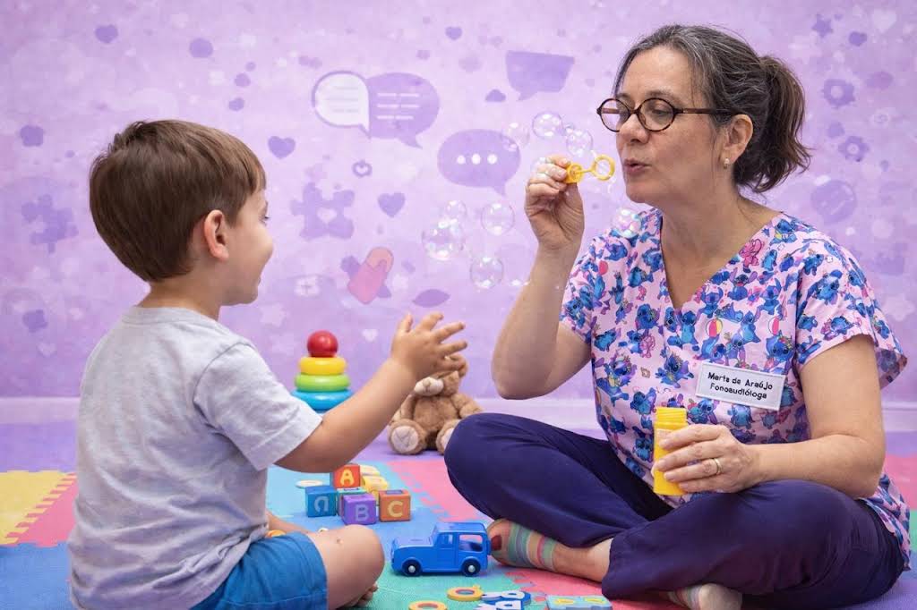 Fonoaudióloga orientando o posicionamento da boca de um menino durante uma sessão de terapia da fala, com espelho e cartões de figuras sobre a mesa.